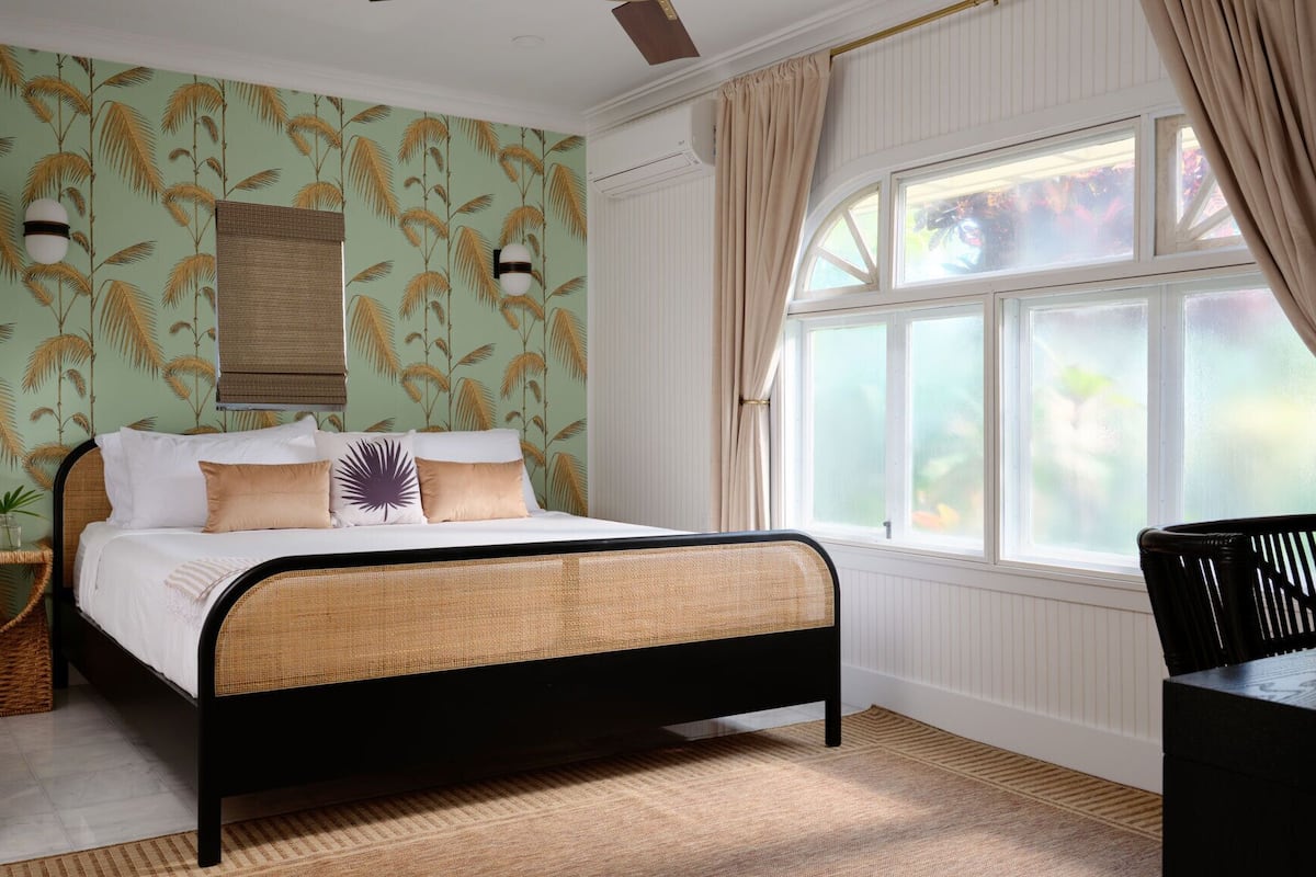 A bedroom with a bed, a window with curtains, a wall with a palm leaf pattern, and a wicker side table.