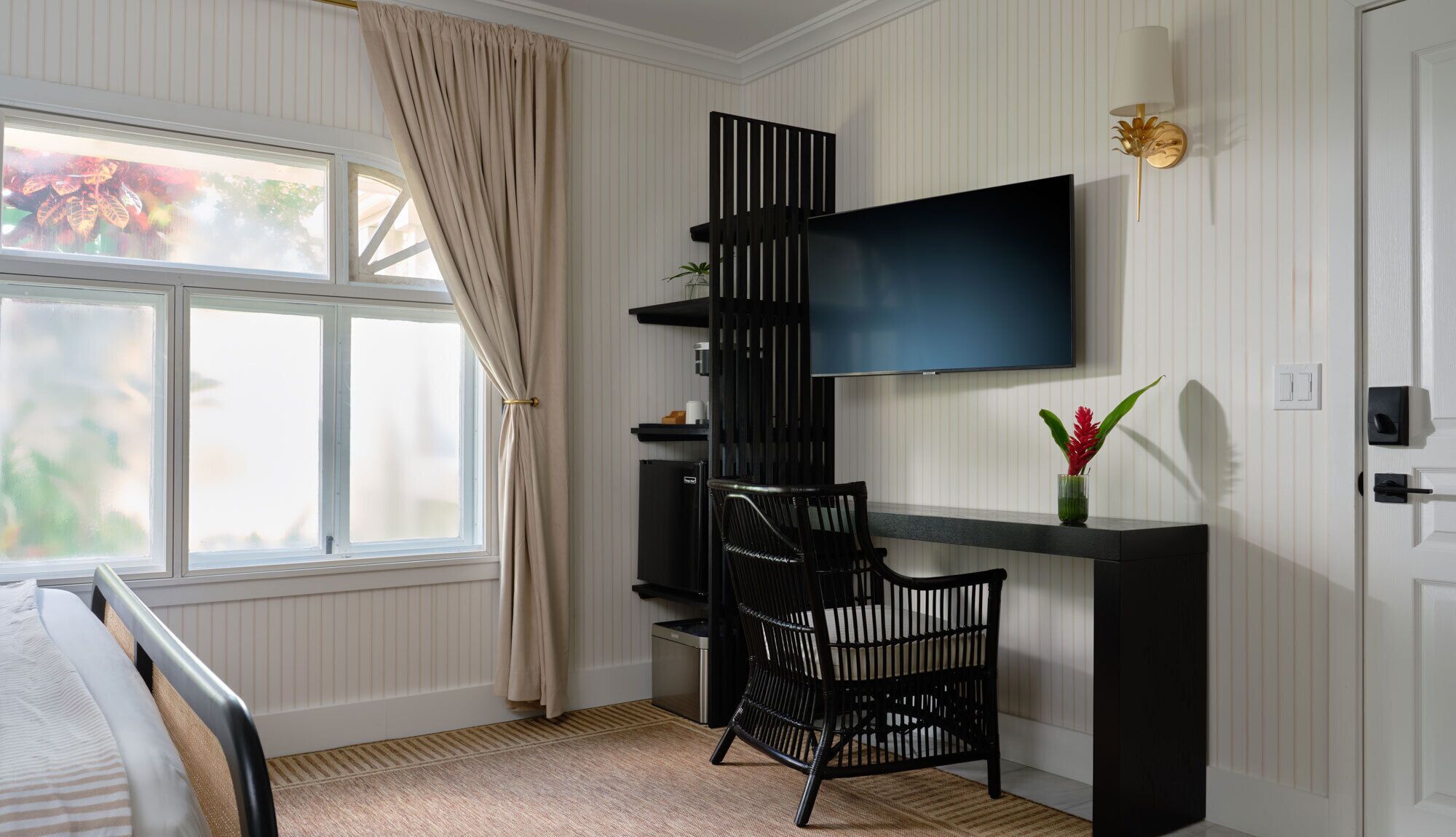 A bedroom with a bed, a TV mounted on the wall, a black chair, a small table, and a window with curtains.