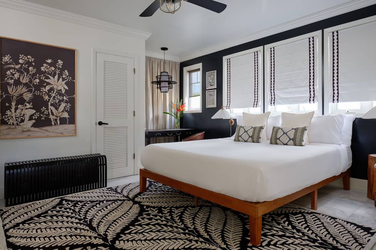 A bedroom with a large bed, a wooden nightstand, a chair, a radiator, a black and white patterned rug, and a ceiling fan.