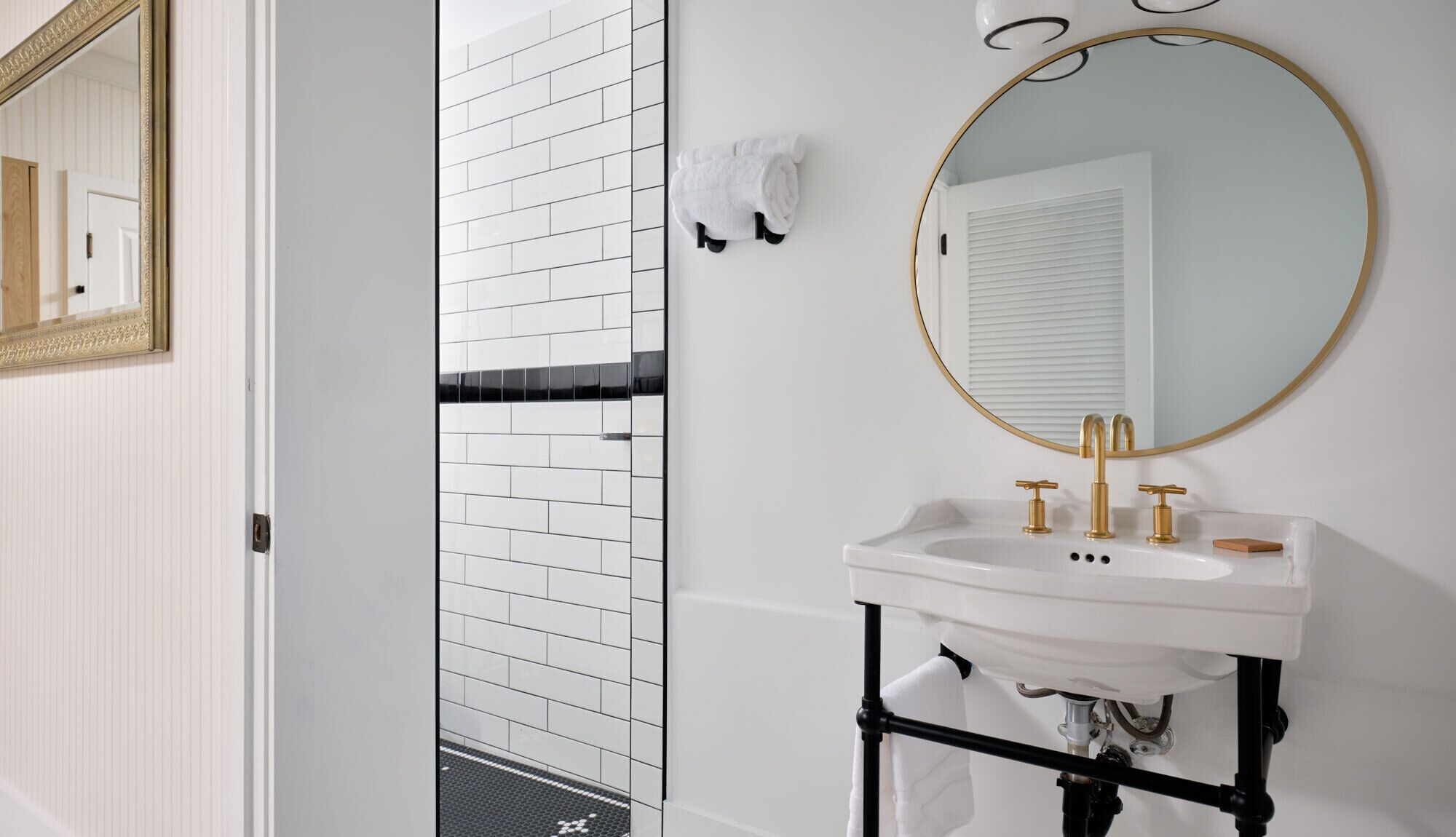A bathroom with a white tiled wall, a black and white tiled shower area, a white sink with gold fixtures, and a round mirror with a gold frame.
