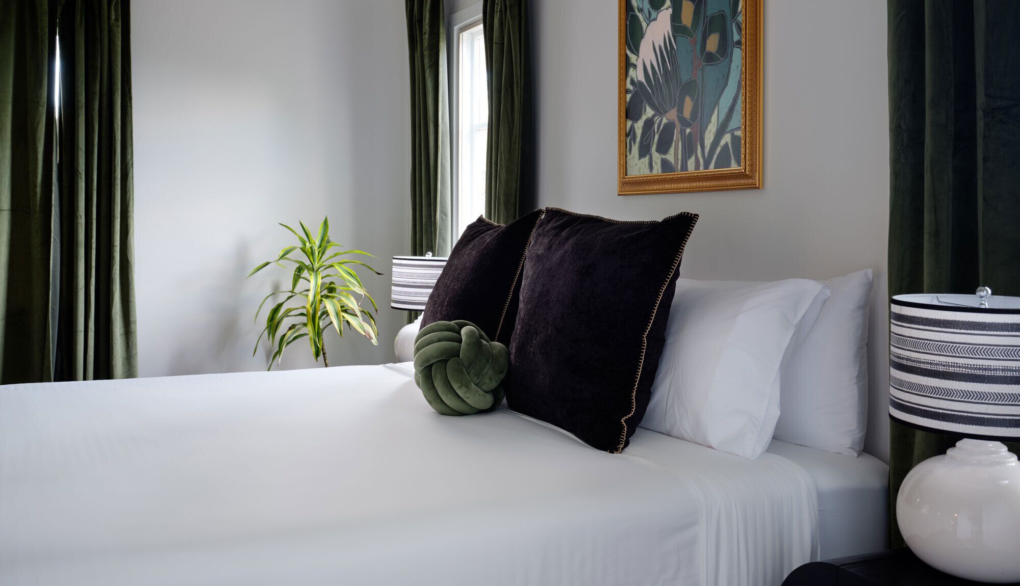 A neatly made bed with a green plant, a dark pillow, and a striped lamp on a bedside table.