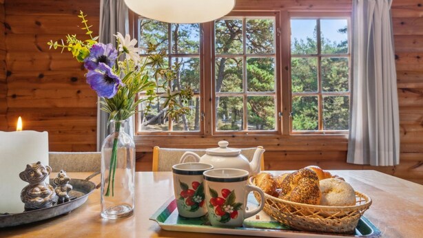 Ein Holzhütten-Interieur mit einem Esstisch, der für ein Frühstück gedeckt ist, darunter Brot, ein Teekessel, Tassen und eine Kerze.