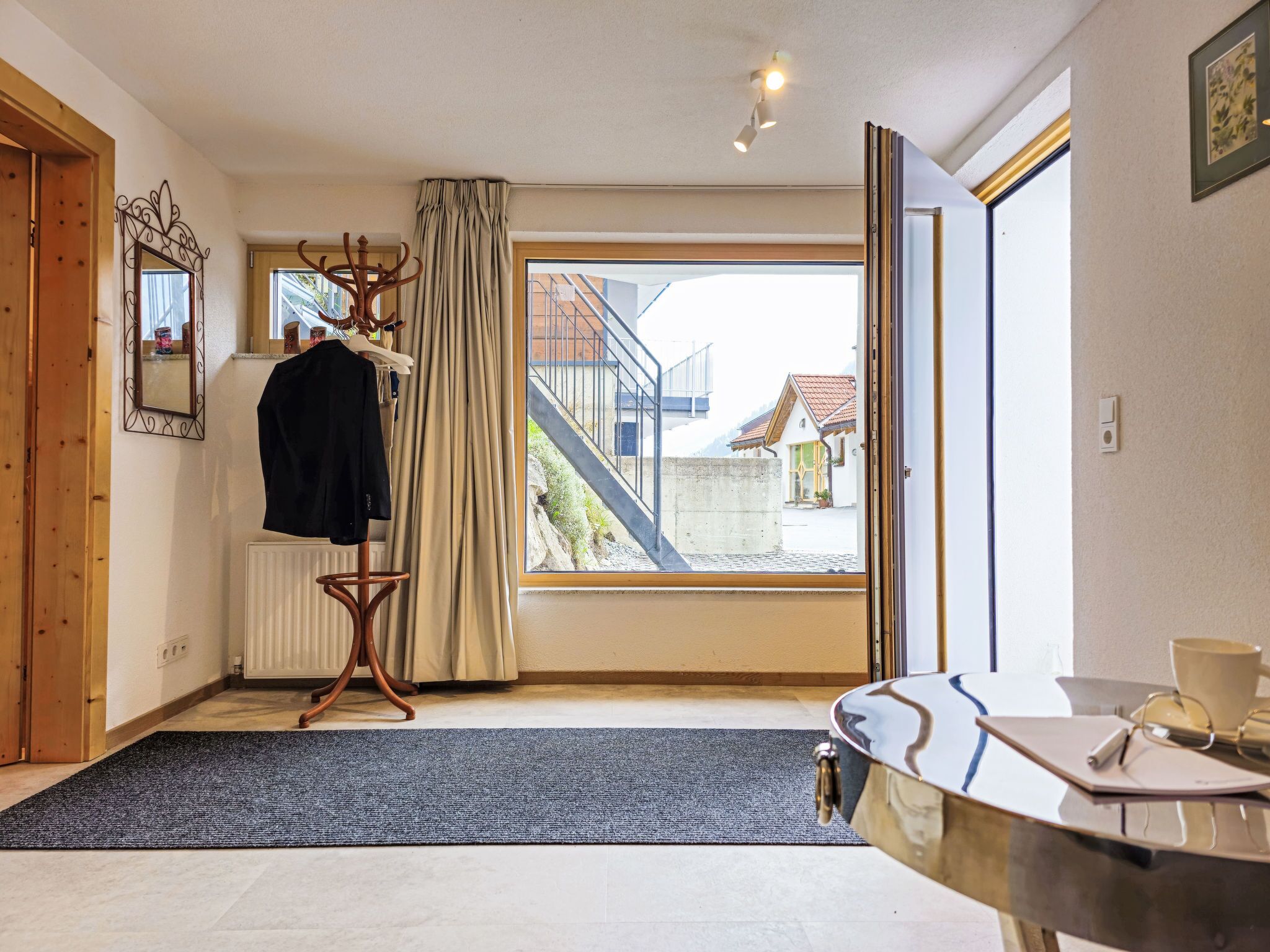 Quarto com uma janela grande, um cabideiro com roupas, um radiador e uma mesa com uma chávena e copos.