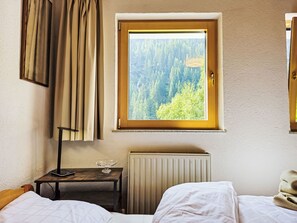 Ein Schlafzimmer mit einem Fenster, das einen Wald zeigt, einem Nachttisch mit einer Lampe und einem Heizkörper.
