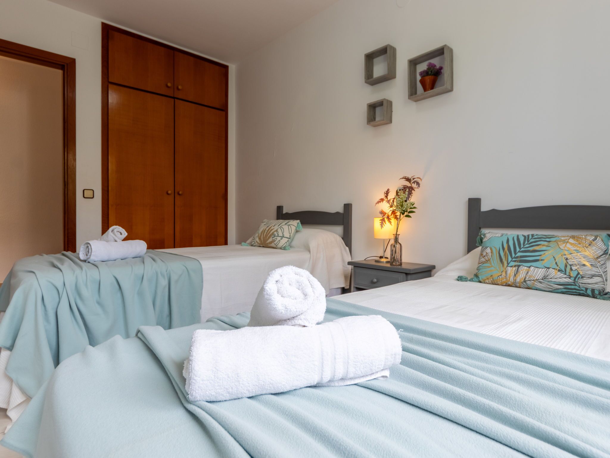 A hotel room with two beds, a wardrobe, a nightstand with a lamp, and a wall-mounted shelf with decorative items.