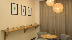 A room with a wooden shelf holding small potted plants, a round wooden table with two chairs, and three framed botanical illustrations on the wall.
