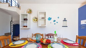Un comedor con una mesa de madera dispuesta para cuatro personas, rodeada de sillas. Hay platos decorativos, una botella de vino y una jarra sobre la mesa.