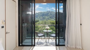 A balcony with a glass door, two chairs, a small round table with a vase of flowers, and a view of a forested hill. - Tropical View Suite A603 at Viva Patong (Patong)