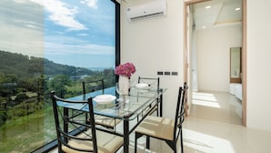 A dining area with a glass table and chairs, a vase with flowers, and a view of a green landscape through large windows. - Tropical View Suite A603 at Viva Patong (Patong)
