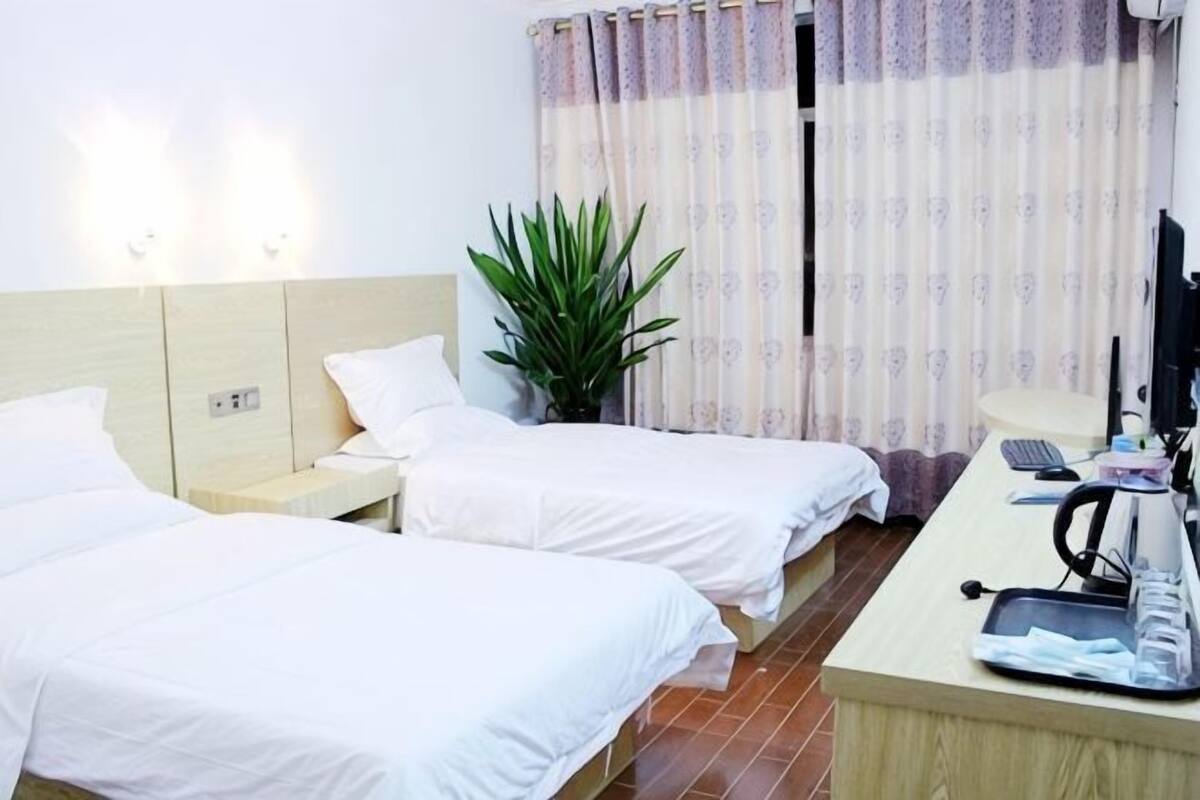 A hotel room with two beds, a desk with a computer, a plant, and a window with curtains.