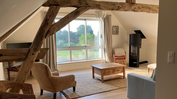 Ein gemütliches Schlafzimmer im Loft-Stil mit freiliegenden Holzbalken, einem großen Fenster, einem Kamin und einer bequemen Sitzecke.
