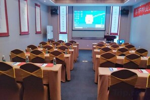 Un salón de conferencias con filas de mesas y sillas, una pantalla de proyector y una pantalla para presentaciones.