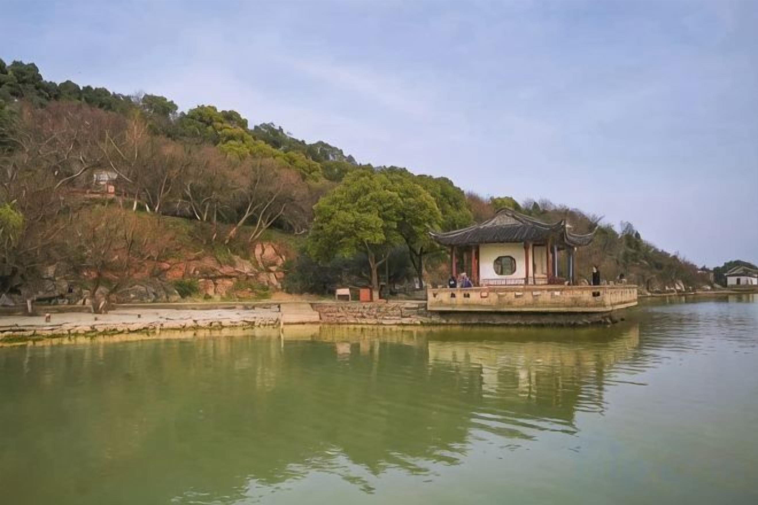 Un pavillon traditionnel situé au bord d’un plan d’eau paisible, entouré d’arbres et de collines.
