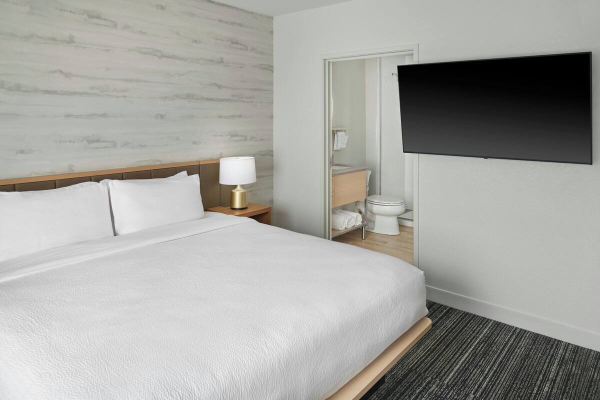 A hotel room with a large bed, a bedside lamp, a flat-screen TV mounted on the wall, and a bathroom visible through an open door.