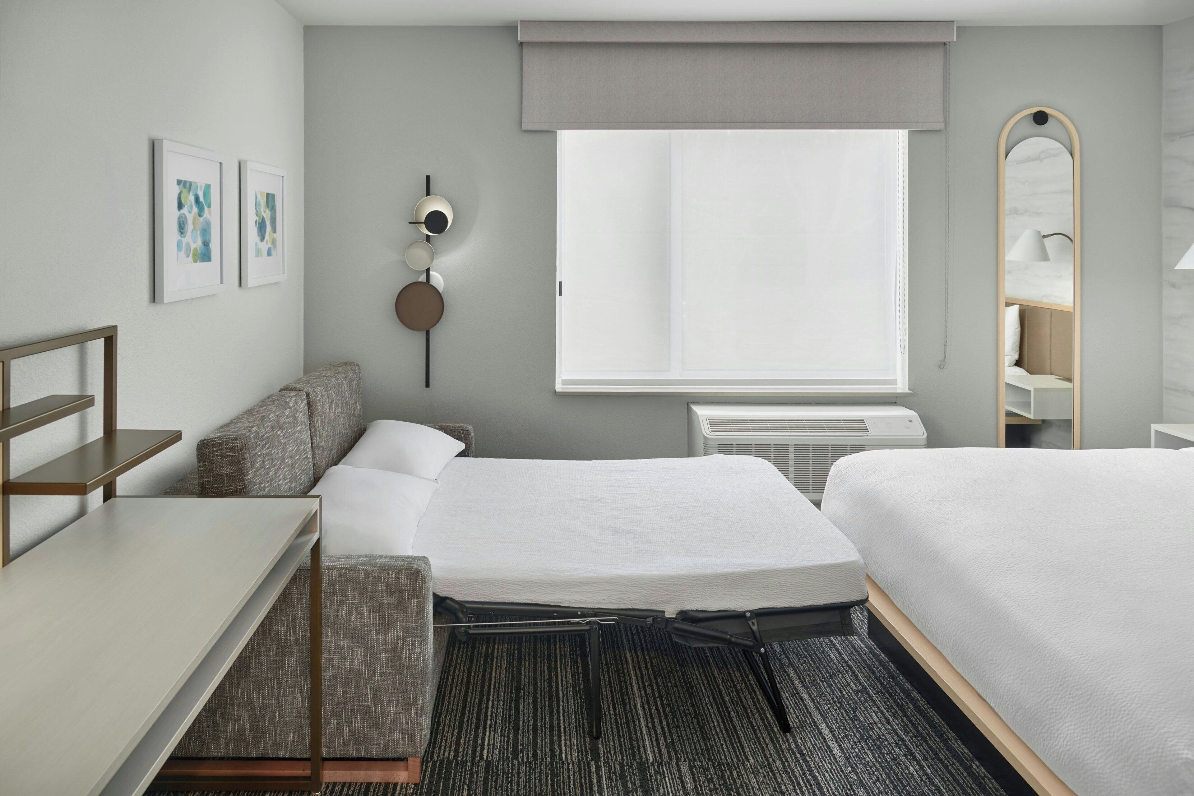 A modern hotel room with two beds, a desk, a chair, a small sofa, a round wall clock, a large mirror, and a window with a view.