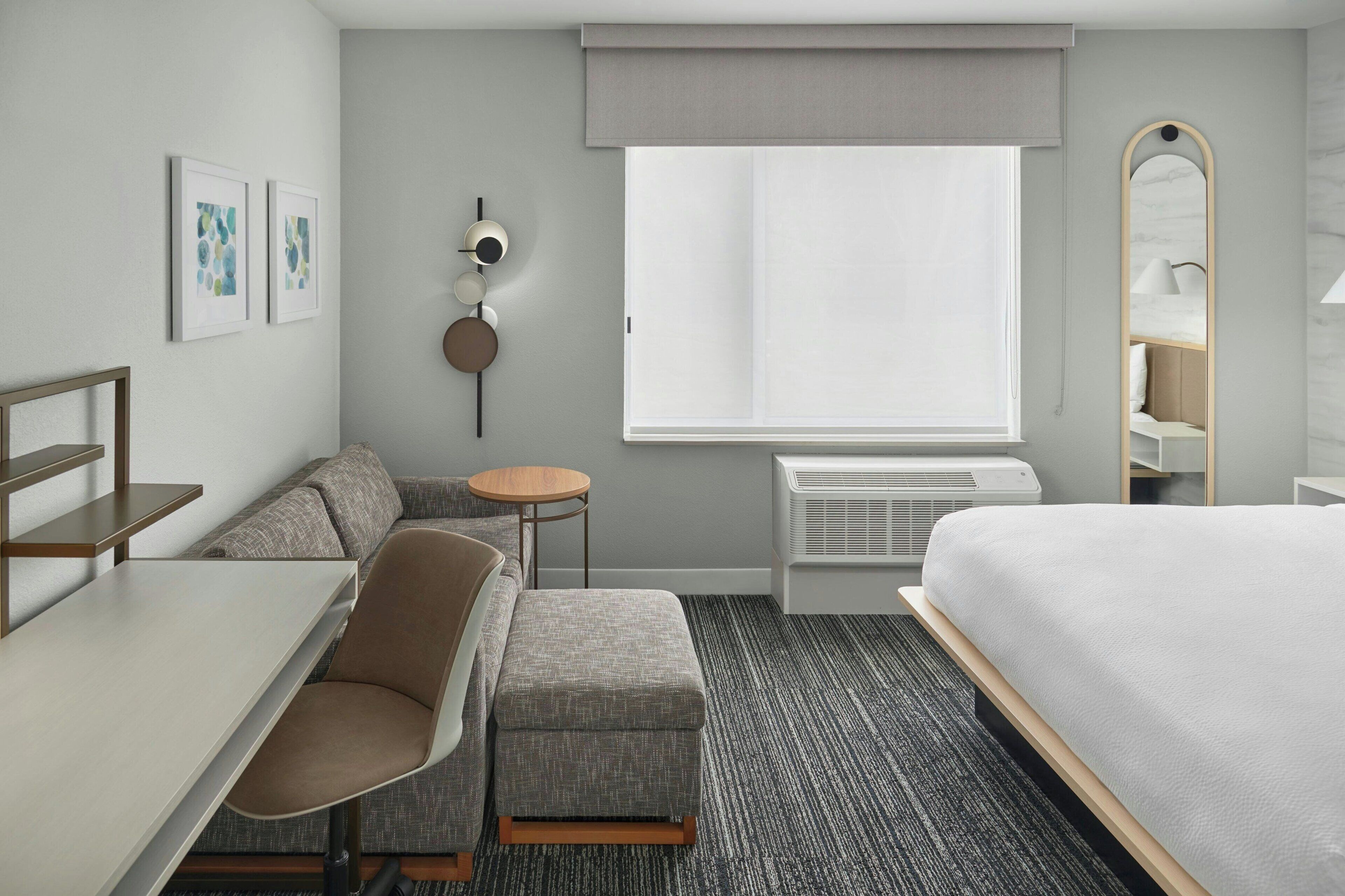Una habitación de hotel moderna con una cama, un escritorio, una silla, una mesita redonda pequeña, un sofá, un espejo y una ventana con vistas.