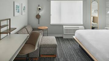 Una habitación de hotel moderna con una cama, un escritorio, una silla, una mesita redonda pequeña, un sofá, un espejo y una ventana con vistas.