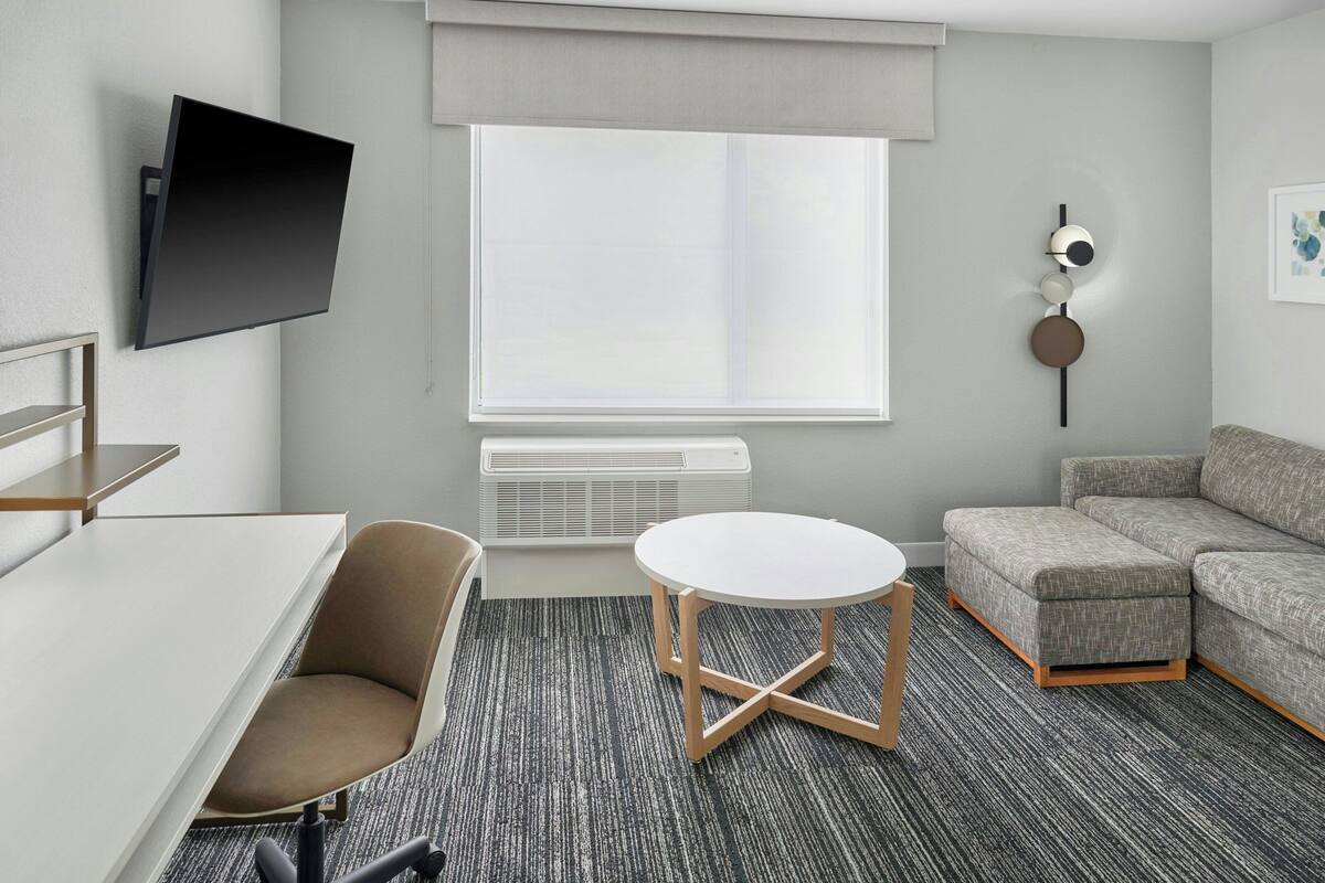 A modern hotel room with a flat-screen TV mounted on the wall, a grey sofa, a round white table, and a grey carpeted floor.