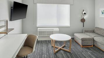 Ett modernt hotellrum med en platt-TV som är monterad på väggen, en grå soffa, ett runt, vitt bord och ett grått, heltäckande golv.