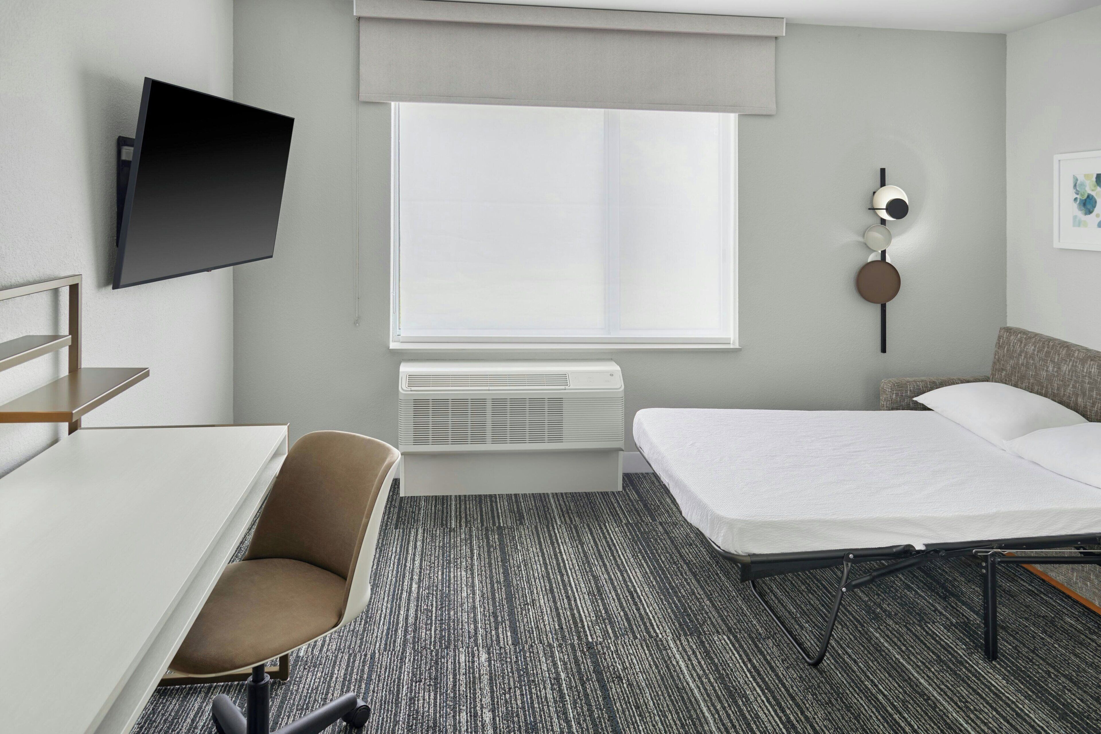 Habitación de hotel con cama, escritorio, silla, televisión de pantalla plana en la pared y ventana con cortina.