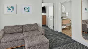 Un moderno cuarto de hotel con un sofá gris, dos cuadros abstractos, un espejo grande y una puerta de madera que da al baño.