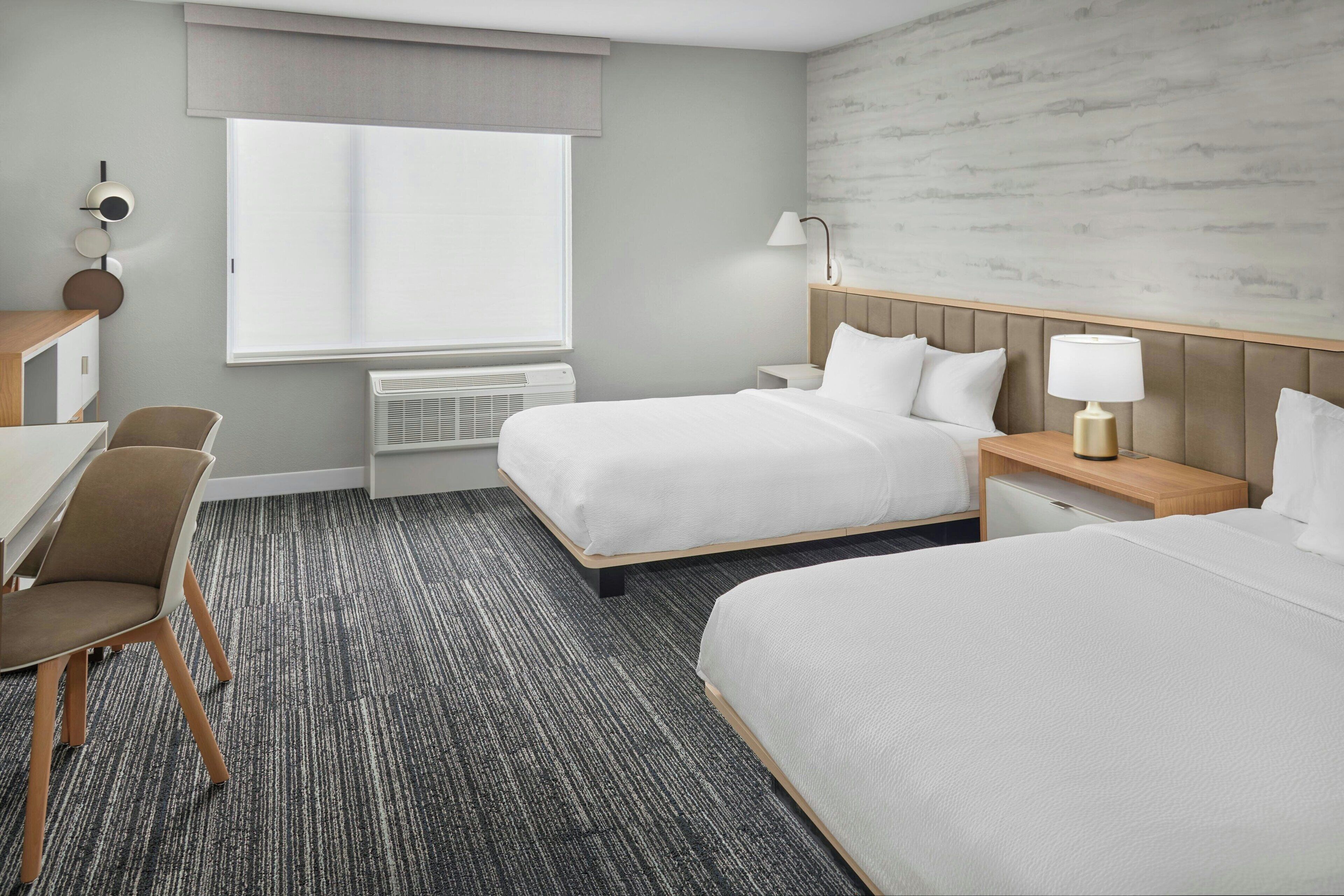 A hotel room with two beds, a desk, a chair, a lamp, and a window with a view of the outside.