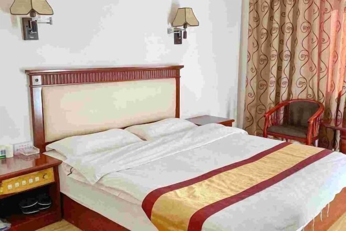 A hotel room with a wooden headboard, a bed with white bedding and a red and gold accent, a nightstand with a lamp, and a chair.