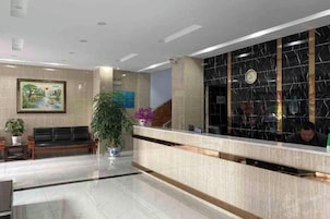 Zona de recepción de hotel con suelo de mármol, mostrador de recepción, área de descanso con sofá y una planta grande en maceta.