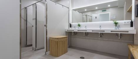 A modern bathroom with a large mirror, three sinks, a shower area, and a trash bin.
