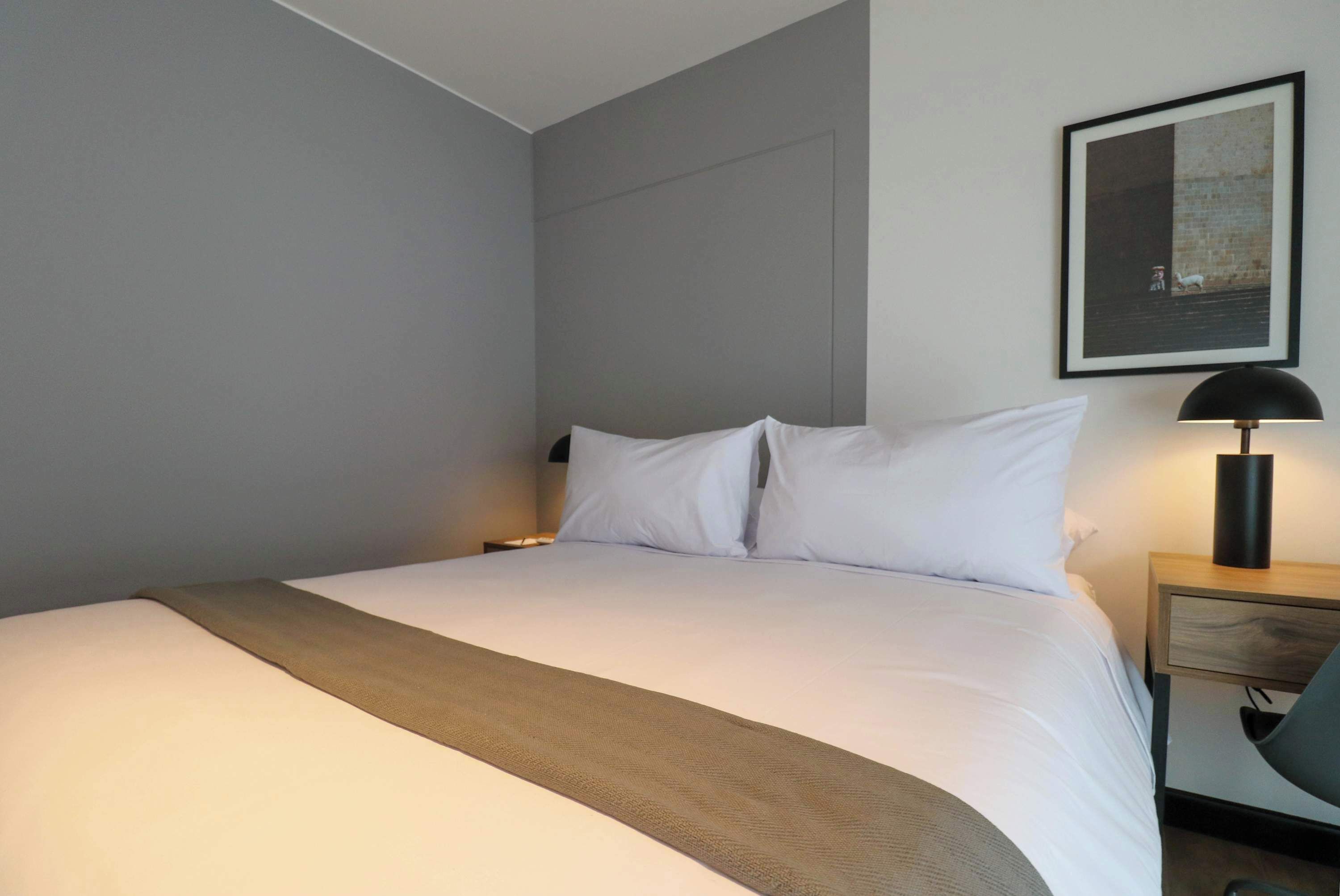 A neatly made bed with white linens and a brown throw blanket, a bedside lamp, and a framed picture on the wall.