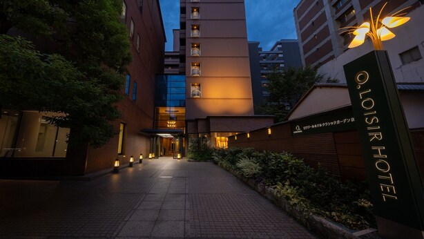 De ingang van Oloisir Hotel met een pad dat naar de ingang leidt, omringd door groen en moderne gebouwen.