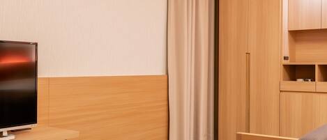 Habitación de hotel con televisor de pantalla plana sobre un escritorio de madera, un reposapiés marrón, un sofá gris y un armario con estantes integrados.
