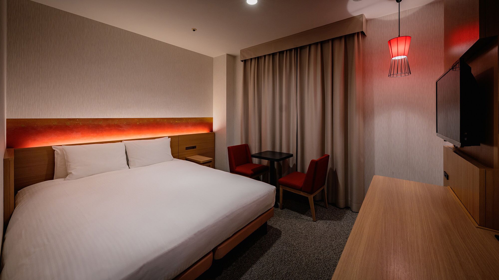 Ein Hotelzimmer mit einem großen Bett, einem Schreibtisch, zwei Stühlen, einem Fernseher und einer roten Pendelleuchte.