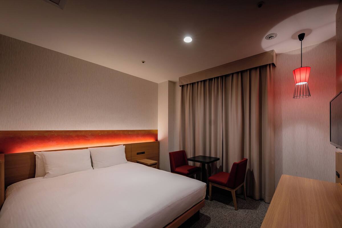 Une chambre d’hôtel avec un grand lit, une tête de lit en bois, une petite table, deux chaises rouges, une télévision et un lampadaire rouge suspendu.