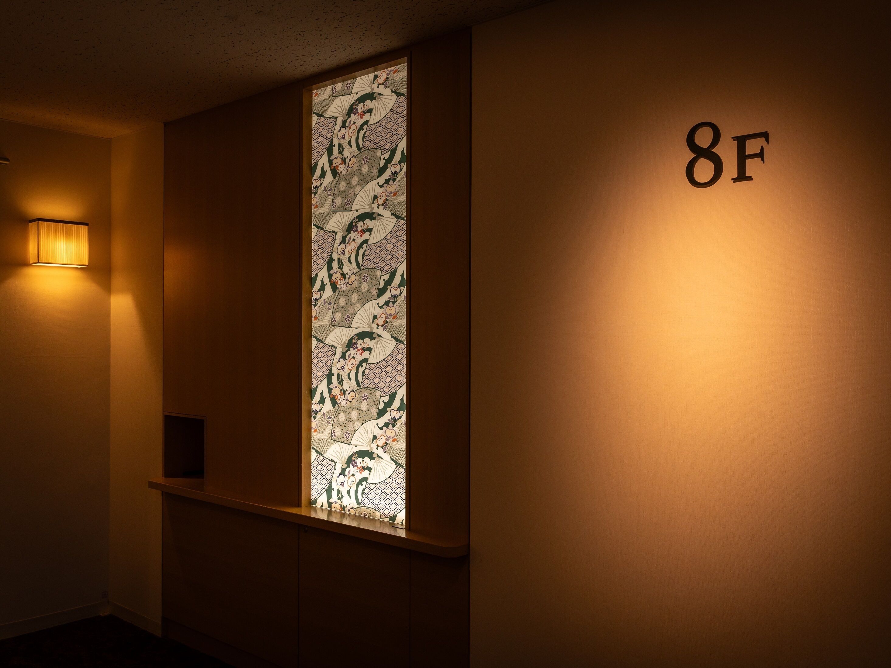 Ein Flur mit einer dekorativen Wandverkleidung in traditionellem Muster und einer beleuchteten Wandleuchte. Auf der Wand ist die Nummer '8F' sichtbar.