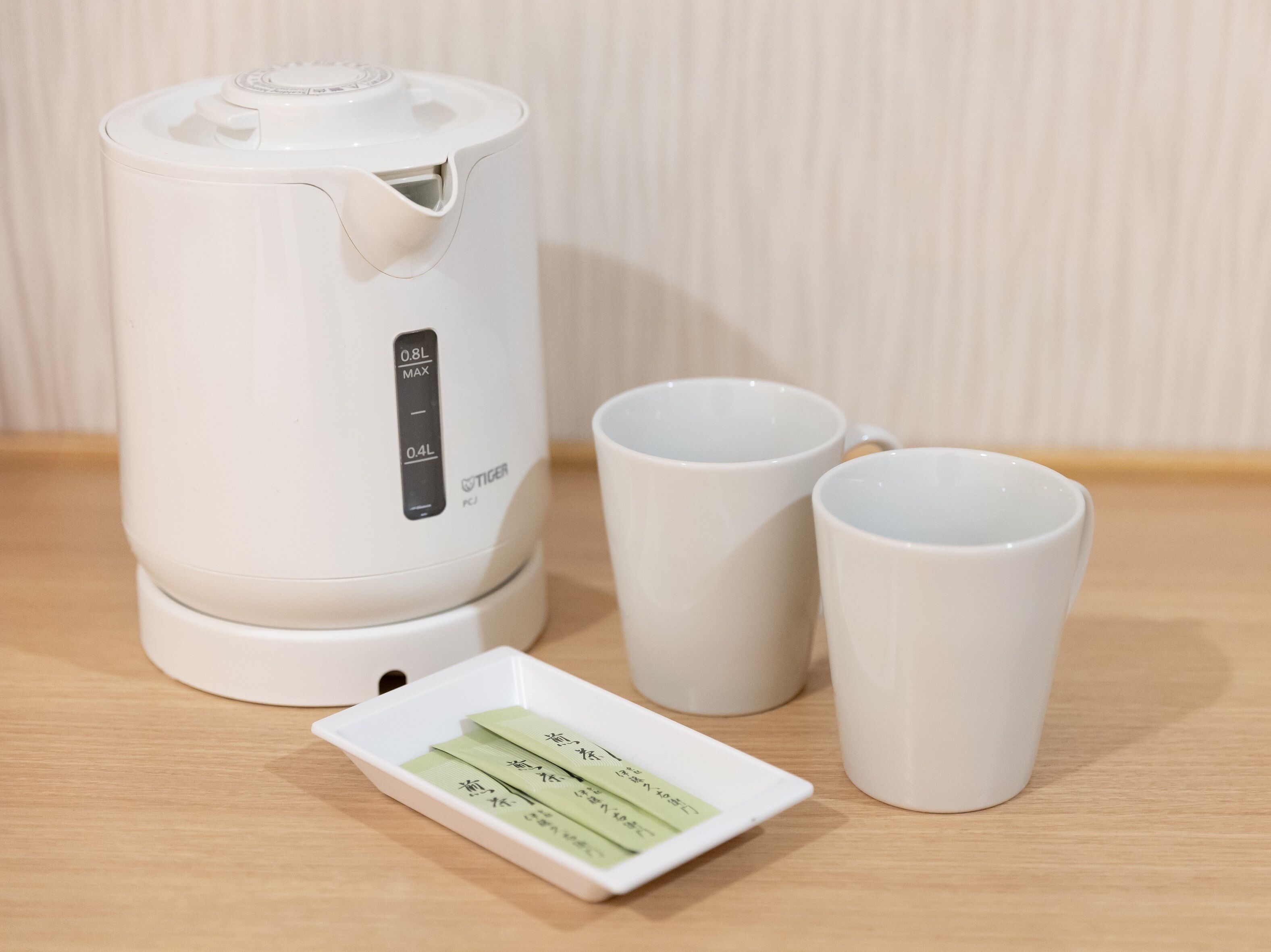 Una tetera eléctrica blanca con pantalla digital, dos tazas blancas y un plato pequeño blanco con bolsitas de té.