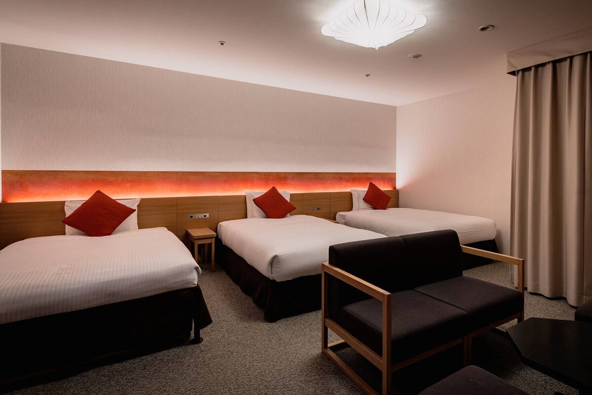 Une chambre d’hôtel avec trois lits, une tête de lit en bois, une petite table et une chaise moderne.