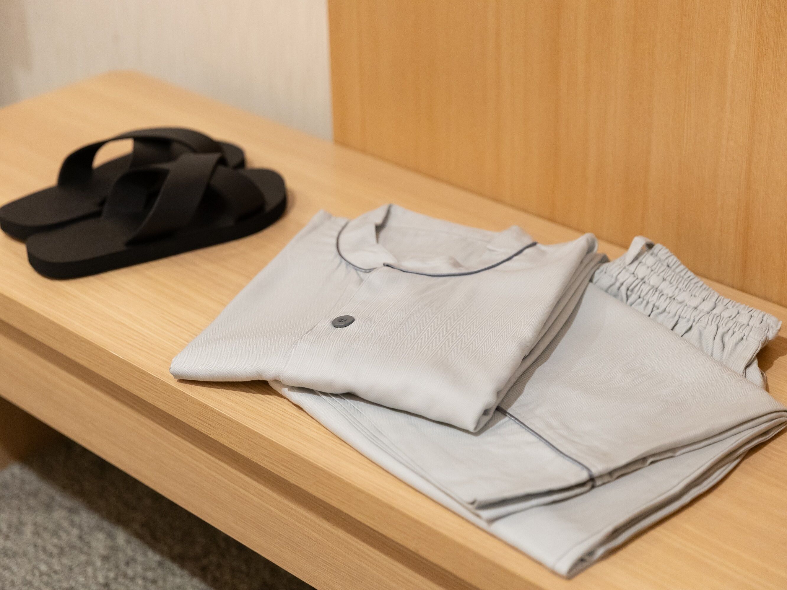 A neatly folded light grey shirt with a button and a black pair of sandals on a wooden bench.