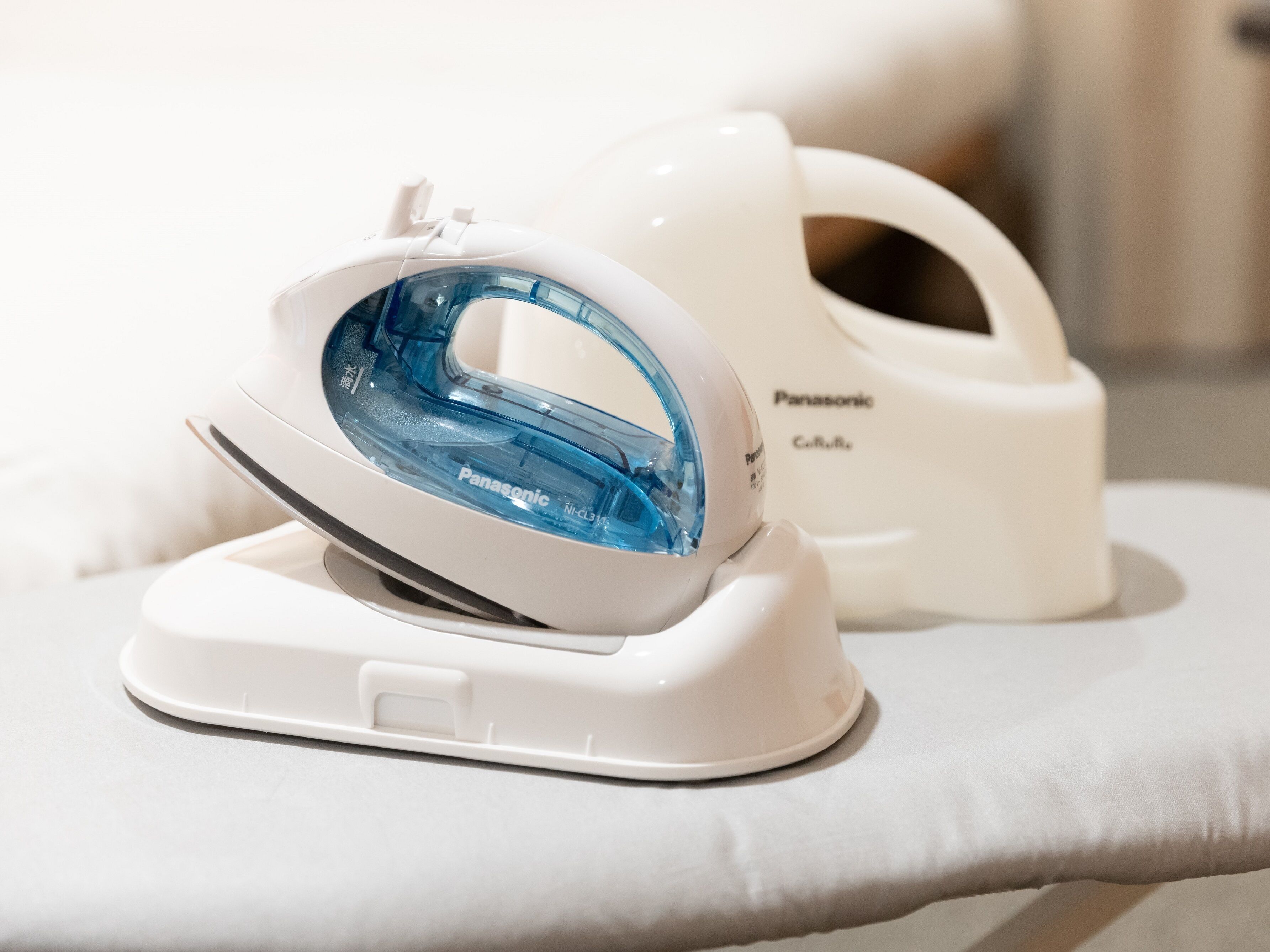 A Panasonic iron with a blue water tank and a white iron, both placed on a white surface.