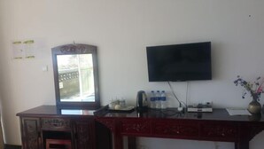 A room with a desk, a television mounted on the wall, a window with a view, and a small table with a kettle and water bottles. - Fushui Inn (Dali City)