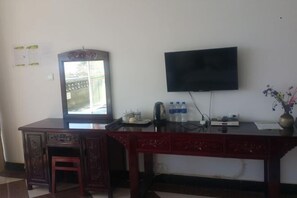 A room with a desk, a television mounted on the wall, a window with a view, and a small table with a kettle and water bottles.