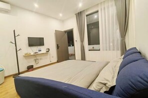 Un dormitorio con una cama, un televisor montado en la pared, una ventana con cortinas y una puerta que da a otra habitación.