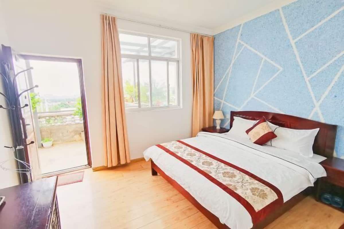 A bedroom with a bed, a nightstand, a lamp, a window with curtains, and a view of greenery outside.