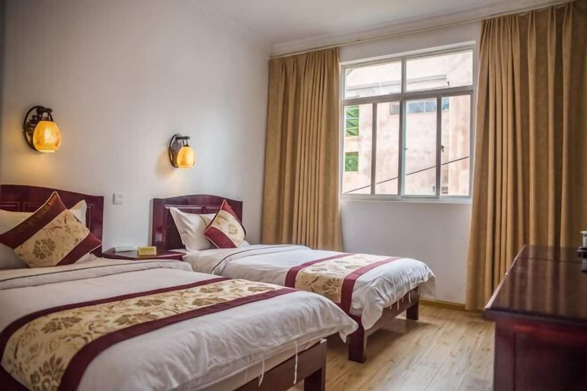 A room with two beds, a window with curtains, a wooden dresser, and wall-mounted lights.