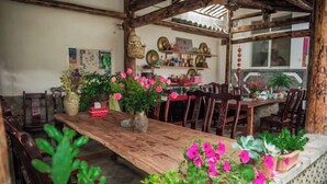 A rustic wooden dining area with a long table, surrounded by wooden chairs. There are potted plants with pink flowers on the table and along the walls. - Fushui Inn (Dali City)