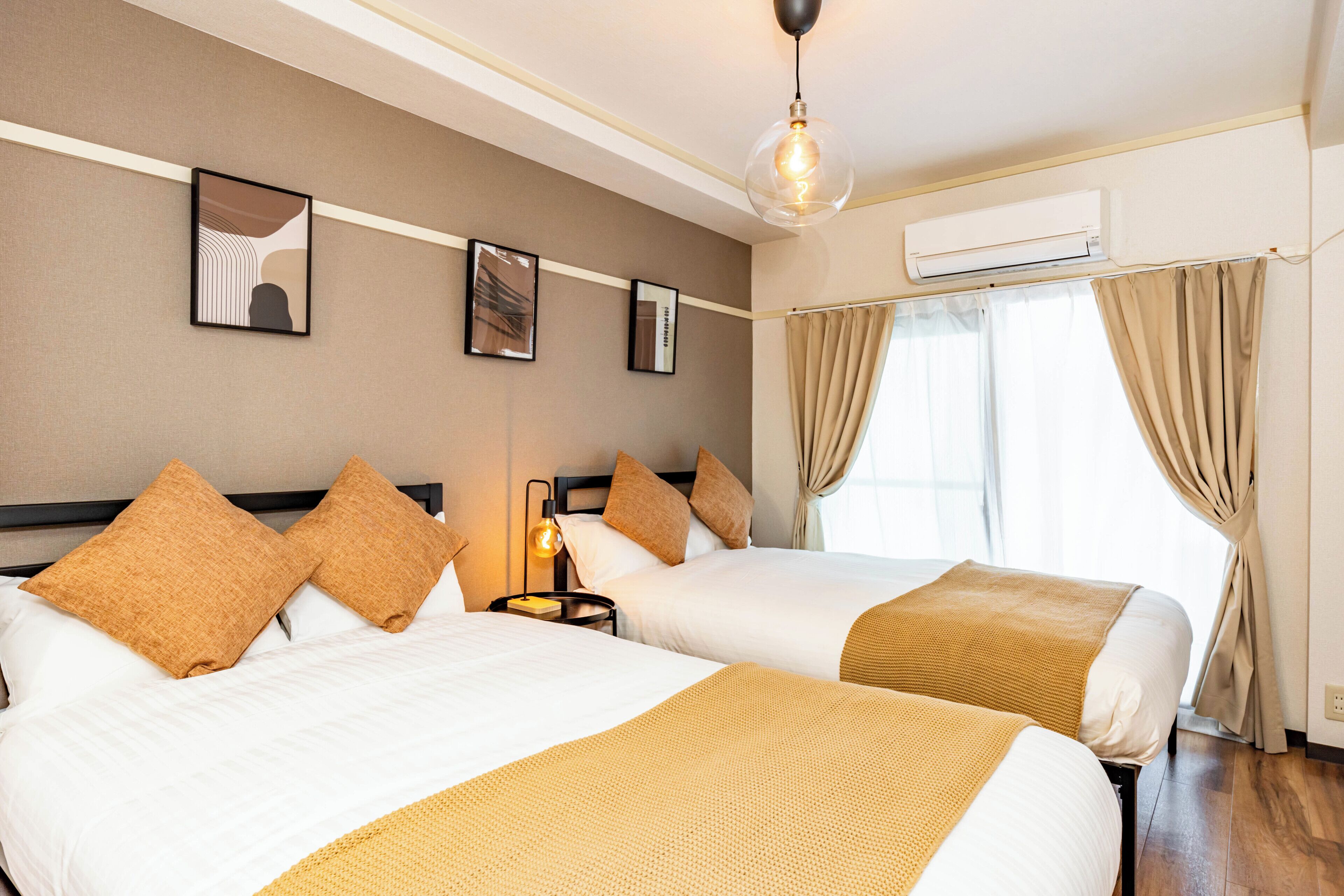 Habitación de hotel con dos camas, cortinas color beige, un equipo de aire acondicionado y cuadros en la pared.