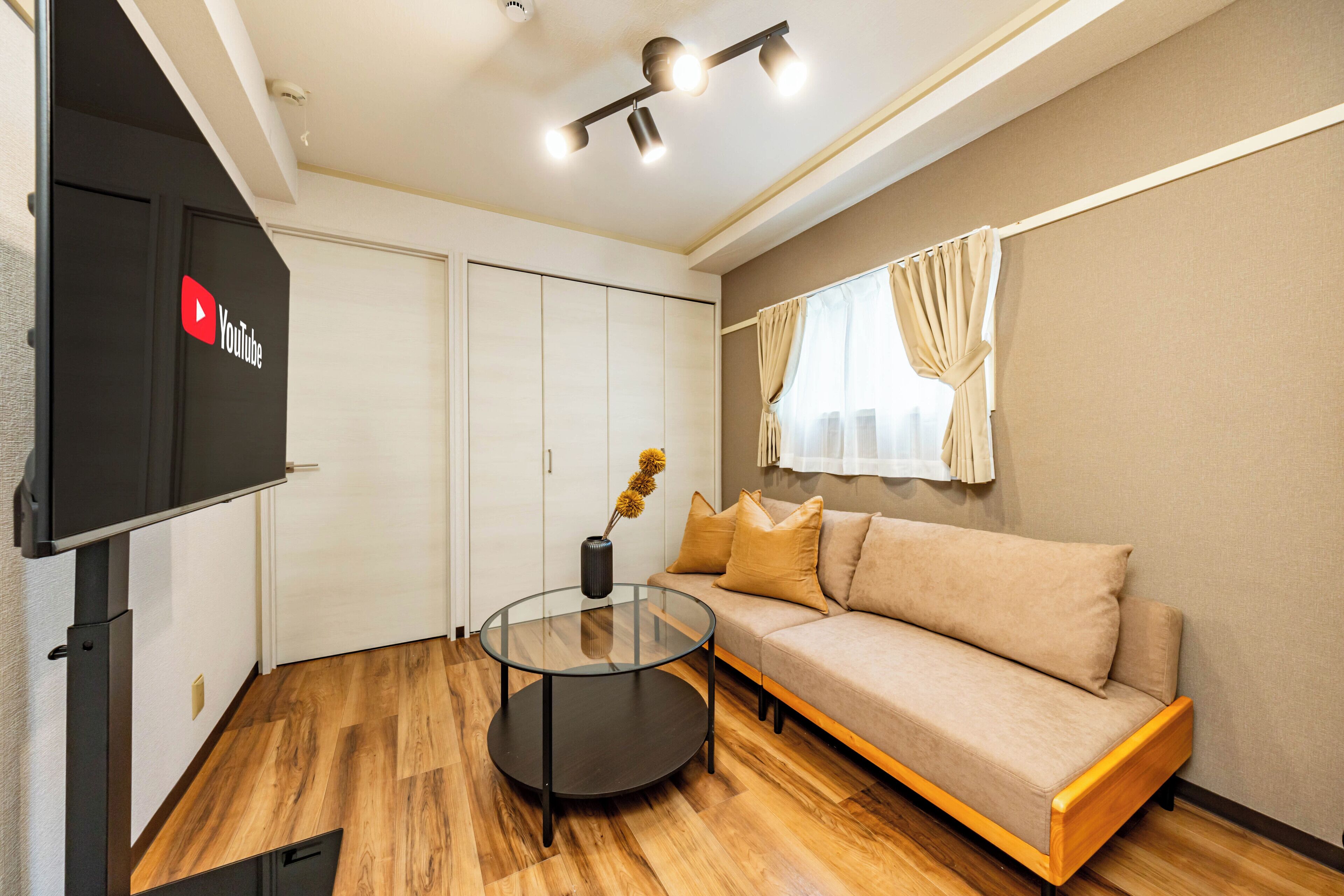 A modern living room with a glass coffee table, beige sofa, wooden flooring, and a flat-screen TV mounted on the wall.