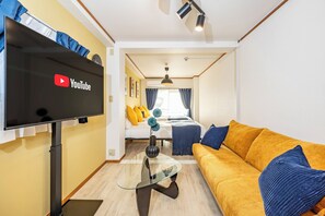 Uma sala moderna com televisão de tela plana, mesa de centro de vidro, sofá amarelo e cama com almofadas amarelas.