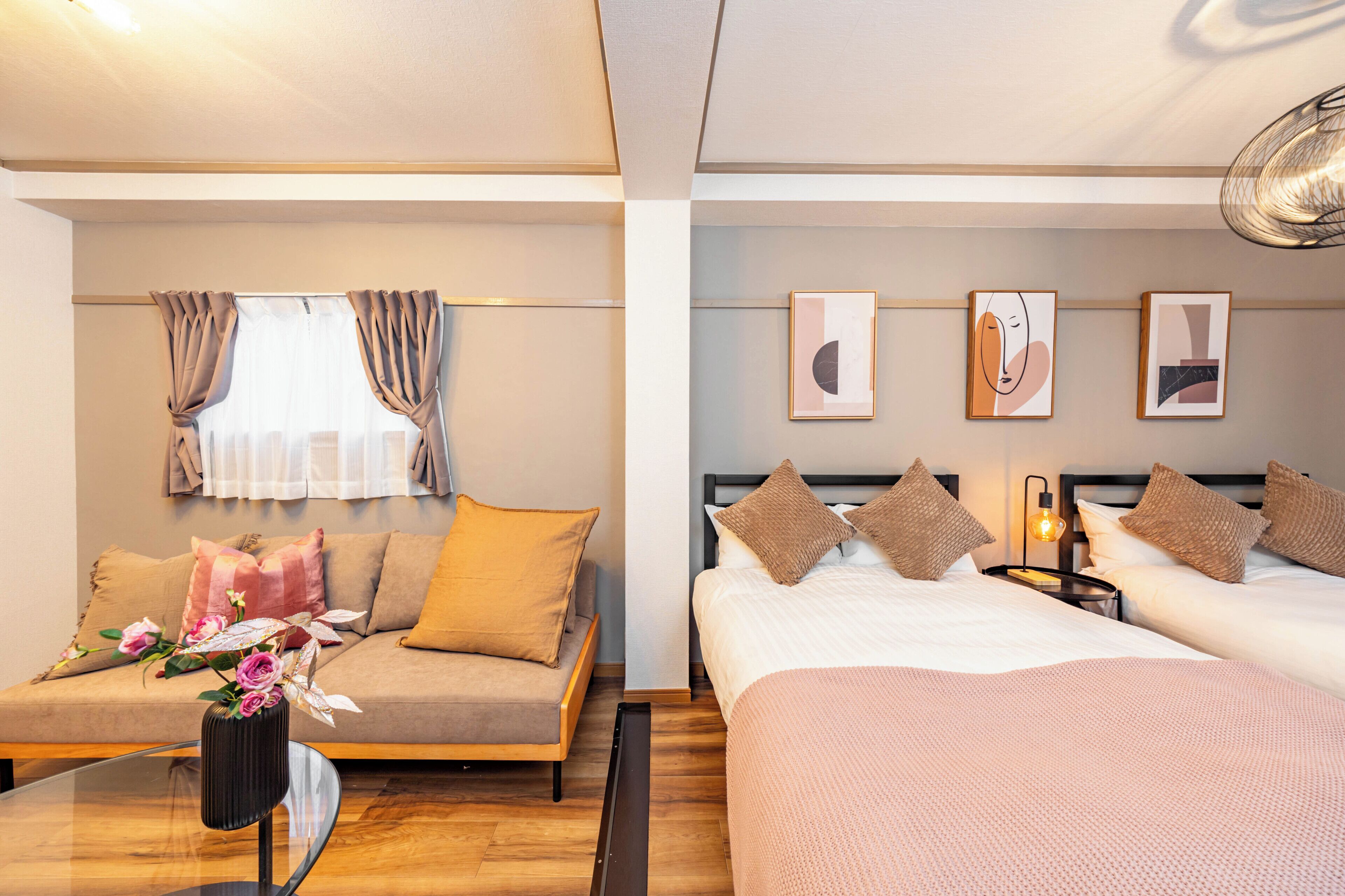 A hotel room with a sofa bed, a separate bed, and a nightstand with a lamp. There are framed artworks on the wall and a window with curtains.