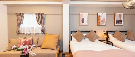 A hotel room with a sofa bed, a separate bed, and a nightstand with a lamp. There are framed artworks on the wall and a window with curtains.
